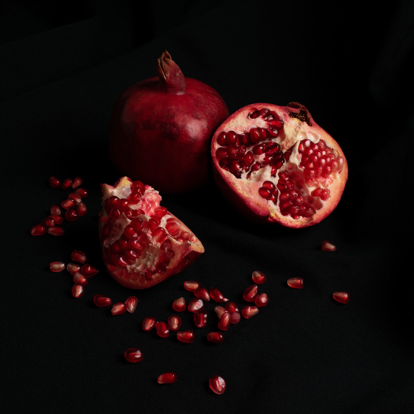 Bright red cut open pomegranate on a black background, with red seeds spilling out around it